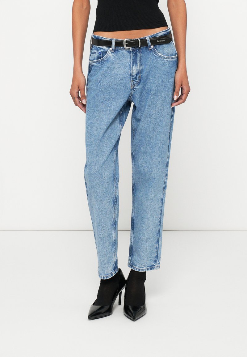 Calças de ganga azul claro com um corte descontraído, com cinto preto, bolsos laterais e ligeiros detalhes desgastados. Combinadas com saltos pretos pontiagudos.