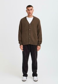 Brown knitted cardigan with a V-neck, six buttons, and ribbed cuffs, paired with black corduroy pants and white sneakers.