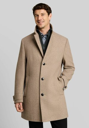 Uomo sorridente che indossa un cappotto beige al ginocchio con bottoni neri sopra un maglione con cerniera e camicia con colletto, mano nella tasca del cappotto.
