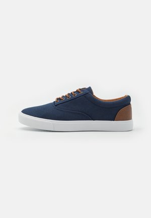 Marine blå canvas sneakers med en hvit gummisåle, brune skinnakenter, runde snører og et strømlinjeformet design.