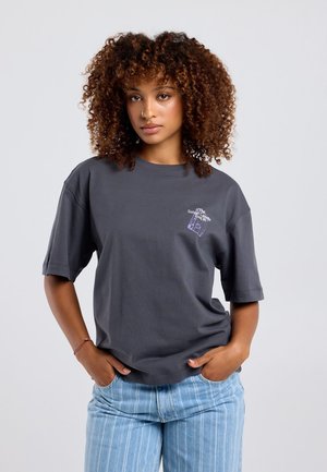 Femme aux cheveux bouclés portant un t-shirt gris foncé avec un petit texte blanc et violet et un jean rayé bleu clair, debout, les mains dans les poches.