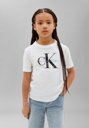 Child with long braided hair wearing a white Calvin Klein t-shirt and light blue jeans, standing against a plain gray background.