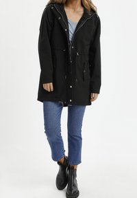 Parka nera impermeabile con cappuccio, zip anteriore e chiusure a scatto. Presenta due tasche frontali e un cordino in vita.