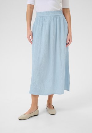 Femme portant une jupe mi-longue texturée bleu clair avec une ceinture élastique, un haut blanc et des chaussures plates beiges, debout sur un fond uni.