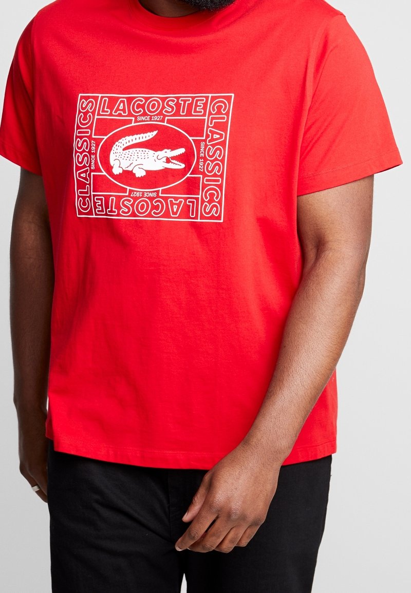 Homme portant un t-shirt Lacoste rouge vif avec un logo crocodile blanc et le texte "Classics Lacoste Depuis 1927", associé à un pantalon noir.