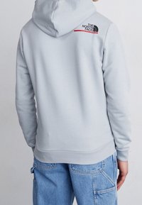 Persona con una sudadera gris claro con el logo de The North Face en la parte superior de la espalda y vaqueros de mezclilla azul con múltiples bolsillos, de espaldas.
