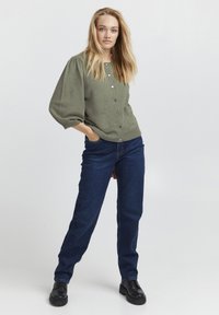 Cardigan in un verde oliva a maglia con maniche a sbuffo, chiusura frontale con bottoni e due tasche, abbinato a jeans a gamba dritta blu scuro e scarpe nere.