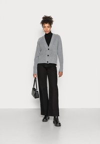 Cardigan cinza de malha com decote em V, com tr�ês botões pretos, utilizado por cima de um suéter de gola alta preto e combinado com calças largas pretas e botas.
