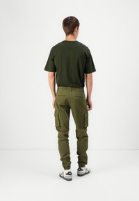 T-shirt in cotone verde oliva e pantaloni cargo. I pantaloni cargo presentano tasche laterali e polsini elasticizzati. Le calzature sono bianche con dettagli neri.