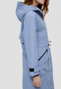 Lichtblauwe waterdichte jas met een capuchon, voorzien van een ritszak, verstelbare trekkoorden en zwarte klittenbandaccenten op de mouwen.