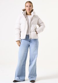 Chaqueta acolchada blanca con capucha y cierre frontal, llevada sobre una camisa blanca, combinada con unos vaqueros de pierna ancha de color azul claro y tacones con estampado.