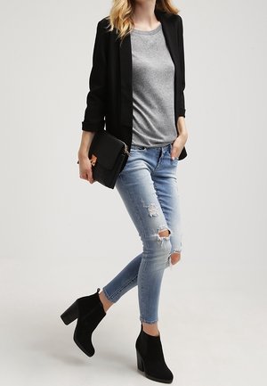 Femme en jean bleu clair déchiré, t-shirt gris, blazer noir, bottines noires, tenant une pochette noire sur un fond uni.