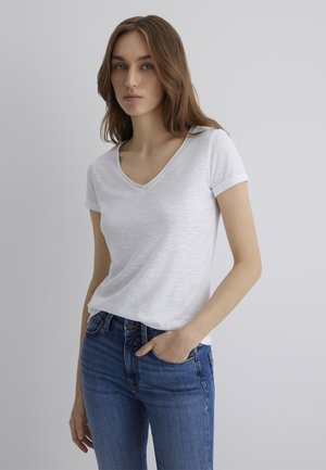 White V-neck t-shirt with short sleeves, made from a lightweight, textured fabric. Paired with blue denim jeans, hands in pocket.