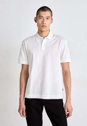 Hombre vestido con una camisa polo blanca de manga corta sencilla y pantalones negros, de pie frente a un fondo claro y uniforme, mirando hacia adelante.