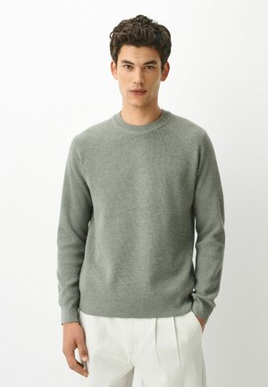 Young man with short curly hair wearing a light gray ribbed sweater and white pants, standing against a plain white background.
