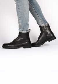 Botas de tobillo de cuero negro con punta afilada, cordones en la parte delantera, cremallera lateral y suela de goma texturizada, llevadas con jeans de un azul claro.