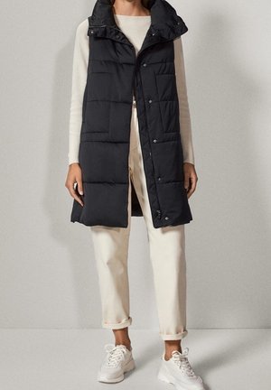Kvinde iført en lang sort vatteret vest over en hvid langærmet top, cremefarvede bukser og hvide sneakers, der står mod en ensfarvet baggrund.