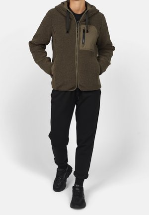 Personne portant une veste à capuche marron duveteuse zippée, un pantalon noir et des baskets noires, debout les mains dans les poches.