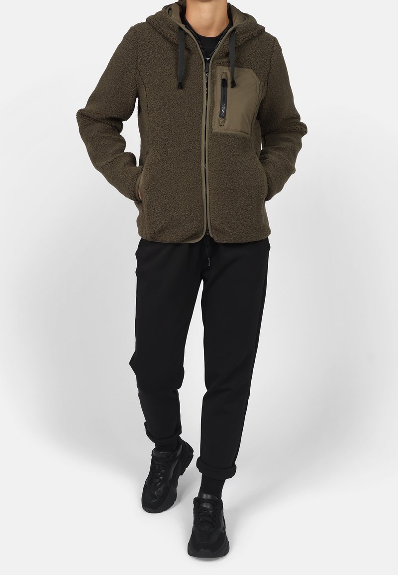 Personne portant une veste à capuche marron duveteuse zippée, un pantalon noir et des baskets noires, debout les mains dans les poches.