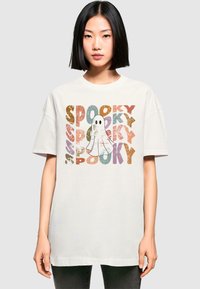 Hvid oversized t-shirt med et legesygt spøgelse design, hvor der står "SPOOKY" i flerfarvede, slidte bogstaver i forskellige størrelser.