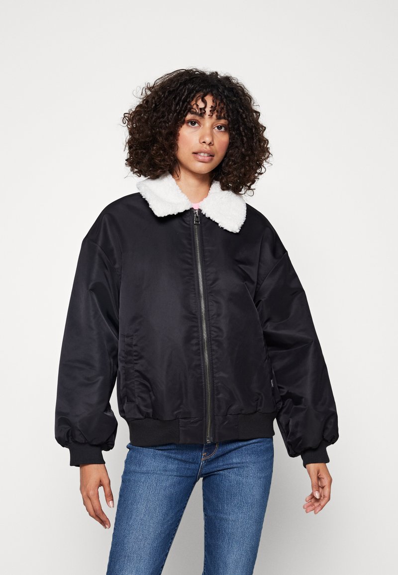Giacca bomber nera con colletto in pelliccia sintetica bianca, chiusura a zip e polsini a costine; presenta una vestibilità leggermente oversize e una texture del tessuto lucida.