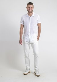 Chemise blanche à manches courtes avec un design à boutons, assortie à un jean blanc et à des baskets blanches avec des semelles marron clair.