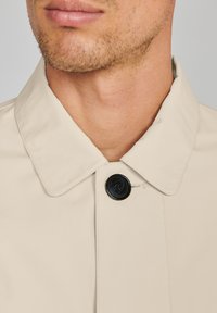Cuello de abrigo beige con un botón negro. Textura suave con detalles de costura visibles. El color crea una apariencia neutral y versátil.