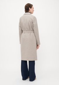 Kvinne som står sidelengs iført en lys beige beltest coat, mørke vidde jeans og beige sko foran en enkel hvit bakgrunn.