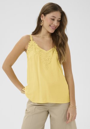 Ung kvinde smilende, iført gul ærmeløs top med blonder, beige bukser, guldringe i ørerne og armbånd, stående med hånden i lommen.