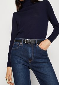 Vrouw in een navy coltrui die in hoge taille blauwe jeans is gestoken, met een zwarte leren riem, hand in de zak en een zilveren ring om haar linkermiddelvinger.