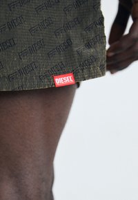 Shorts en tissu vert arborant un motif en relief subtil "DIESEL", avec une étiquette logo rouge à l'ourlet. La texture semble légèrement froissée.