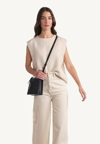 Top en tricot sans manches beige, col montant, associé à un pantalon large crème ; le modèle tient un petit sac noir avec une longue bandoulière.
