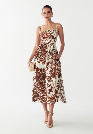 Frau trägt ein ärmelloses, braun-weiß geblümtes Midikleid mit dünnen Trägern, hält eine kleine beige Clutch und trägt goldene Creolen.