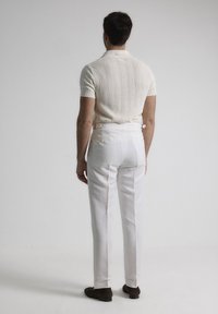Camisa polo de manga corta de punto en color crema, combinada con pantalones blancos ajustados que presentan detalles de hebilla lateral, vista desde la parte posterior.