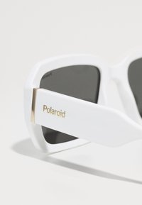 Lunettes de soleil à monture carrée blanche avec des verres sombres, accents dorés sur les branches et logo Polaroid sur le côté. Texture en plastique lisse.