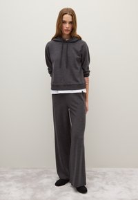 Felpa grigia con cappuccio e polsini a coste, abbinata a pantaloni larghi coordinati. Texture liscia, indossata sopra una camicia bianca, portata con pantofole nere.