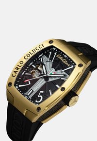 Goudkleurig vierkant horloge met een zwart gestructureerd wijzerplaat, witte uurmarkeringen en een zwarte rubberen band. Beschikt over een zichtbare beweging en rode accenten.