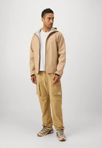 Beige Outdoor-Jacke mit grauem Futter, kombiniert mit hellen, sandfarbenen Cargohosen mit mehreren Taschen. Graue und beige Schuhe vervollständigen den Look.