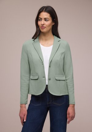 Femme portant un blazer texturé vert clair sur une chemise blanche et un jean bleu foncé, debout devant un fond beige uni.