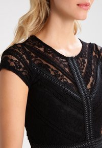 Robe en dentelle noire à manches courtes, présentant un motif texturé, un décolleté en V, et des détails tressés complexes le long des coutures.