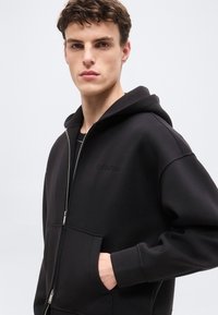 Νεαρός άνδρας που φοράει μαύρο φούτερ με φερμουάρ και κουκούλα, χέρι στην τσέπη, κοιτάζοντας ελαφρώς προς τα πλάγια μπροστά σε απλό ανοιχτό φόντο.