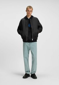 Veste bombardier noire avec une fermeture éclair argentée, portée par-dessus un sweat à capuche noir avec des graphismes bleus, associée à un jean en denim clair et des chaussures noires.