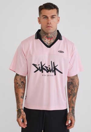 Mand med tatoveringer på nakke og arm iført en pink SikSilk trøje med sort krave og teksten "Ride or Die", stående foran en ensfarvet baggrund.