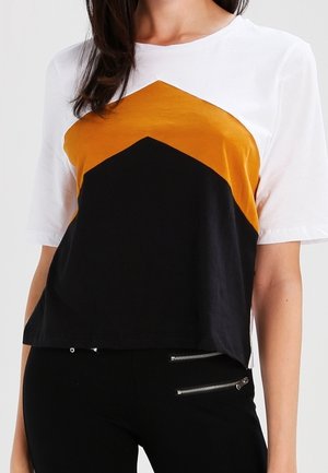 Kobieta ubrana w biały top z krótkim rękawem ozdobiony wzorem w musztardowo-czarne jodełki, w połączeniu z czarnymi spodniami z dwoma kieszeniami na zamek.