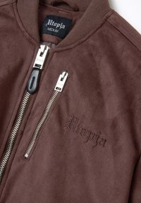 Giacca bomber in suede marrone con zipper argentato, caratterizzata da un'etichetta del marchio nera e argentata e la scritta ricamata "Utopia" in marrone abbinato.