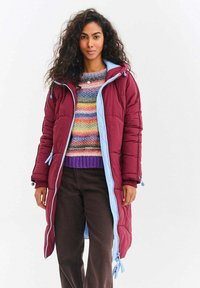 Bordový péřový kabát s světle modrou podšívkou, se zipem a nastavitelnými stahovacími šňůrkami. Nosený přes vícebarevný pruhovaný svetr a tmavé kalhoty.