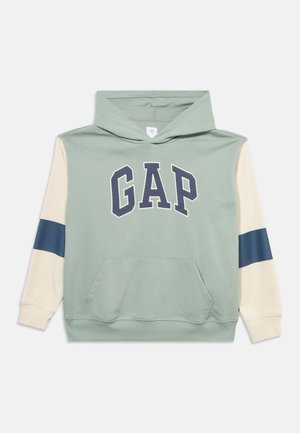 Kapucar z zelenim svetlim telesom, kremnimi rokavi z mornarsko modrimi črtami, sprednjim žepom v obliki kenguruja in logotipom "GAP" v mornarsko modri barvi na prsih.