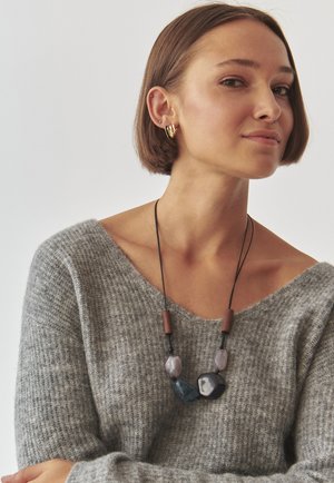 Maglione grigio a maglia con scollatura ampia. La collana presenta perle irregolari in tonalità di grigio, blu e nero, infilate su un cordino nero con dettagli in legno.
