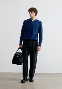 Άνδρας που φοράει μπλε πόλο με μακριά μανίκια, μαύρο παντελόνι και μαύρα παπούτσια, κρατώντας μια μαύρη δερμάτινη τσάντα duffel, στέκεται σε απλό φόντο.