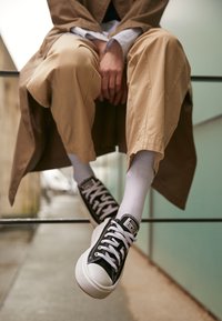 Zwarte canvas sneakers met witte rubberen zolen, witte veters en zwarte accenten. Het model draagt lichtgekleurde broek en witte sokken.